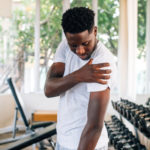 Side view of muscular African American man standing and suffering from shoulder pain during workout with dumbbells. Sportsman holding sore shoulder in gym