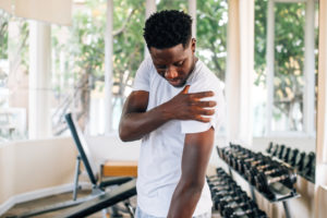 Side view of muscular African American man standing and suffering from shoulder pain during workout with dumbbells. Sportsman holding sore shoulder in gym